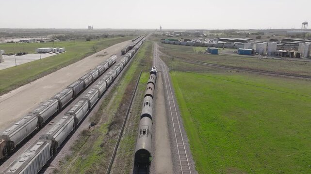Aerial tracking shot of an oil train (tank cars) pulling out of an industrial rail yard past freight cars on a sunny day, showcasing energy logistics.