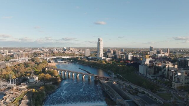 Aerial descent over Mississippi River, Minneapolis. Features Stone Arch Bridge, St. Anthony Falls Dam, and Downtown Skyline with the Eleven Tower and Gold Medal Flour sign at sunset golden hour.