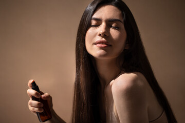 Young woman with long dark hair is applying fragrance from a spray bottle, enjoying the scent in a soft-lit environment, capturing a moment of personal care and beauty