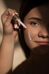 Obraz premium Young teen girl applying skincare serum to her face with a dropper, showcasing a beauty routine in soft lighting, highlighting skin texture and natural beauty