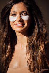 Young woman with long hair wearing under-eye patches smiles warmly, showcasing skincare routine and natural beauty in a soft-lit environment, emphasizing self-care and wellness