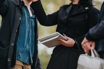 A group of young professionals meet outdoors, sharing ideas while one person holds a tablet. They wear smart casual outfits, suggesting a collaborative moment in a business or networking setting.