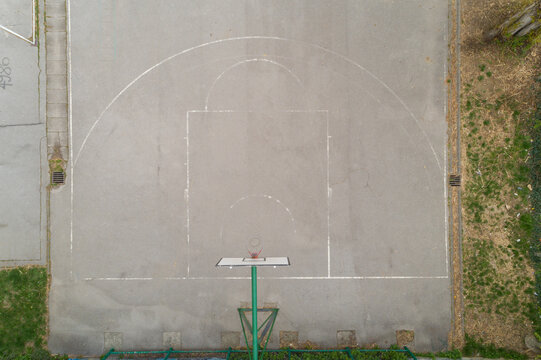Aerial view of the stark concrete of a basketball court, its faded white lines a ghostly echo of games played, contrasting with the encroaching green vegetation, Zagreb, Croatia.