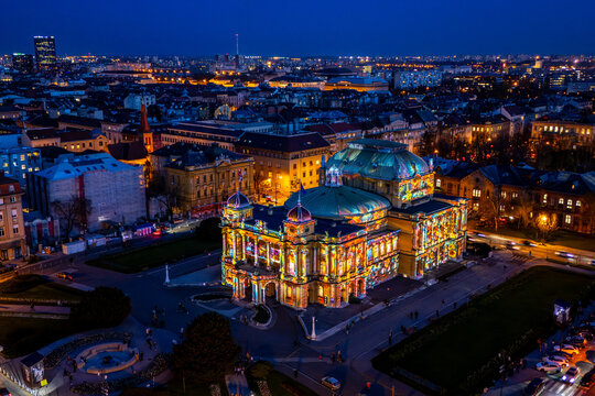 Aerial view of the Croatian National Theatre bursts with vibrant light against the darkening cityscape, a beacon of culture amid the urban sprawl, Zagreb, Croatia.