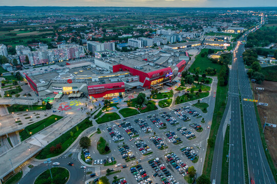 Zagreb, Croatia - 18 November 2025: Aerial view of the vibrant Arena Centar, a modern shopping complex, glows with red accents against a backdrop of orderly parking lots and verdant city surroundings.