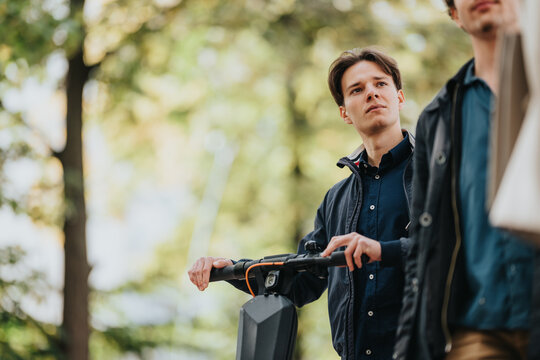 Two young colleagues in casual business attire pause outdoors beside a modern electric scooter in a sunny park, suggesting flexible commuting and a contemporary urban lifestyle.