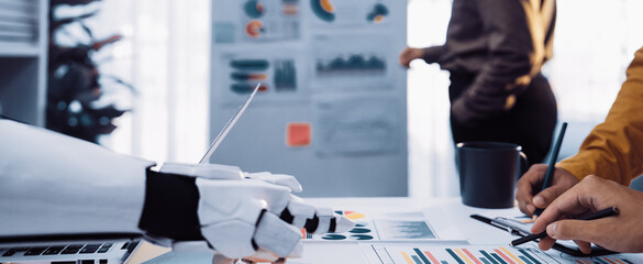 A close-up view of a workspace featuring a robotic arm and human hands working on charts and graphs, illustrating collaboration in a modern office environment.Noogenesis