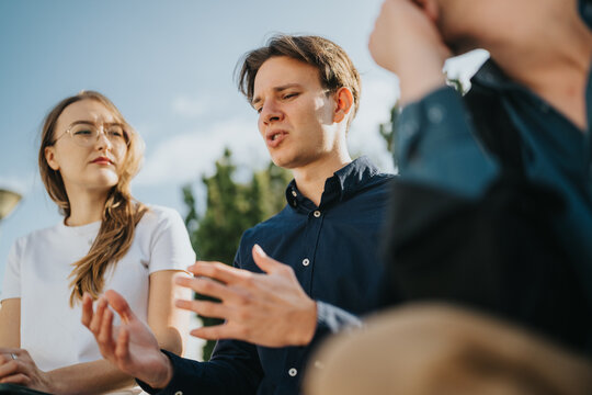 A group of young colleagues chat and gesture while outside, focused on a discussion. The scene conveys teamwork, collaboration, and friendly professional interaction in a sunny, open-air setting. - Powered by Adobe