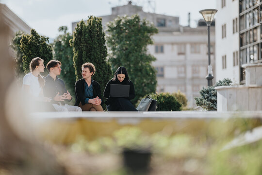 A group of colleagues sit on a bench in a sunny urban park, discussing ideas while one person uses a laptop. The scene conveys collaboration, casual professionalism, and a productive outdoor vibe.