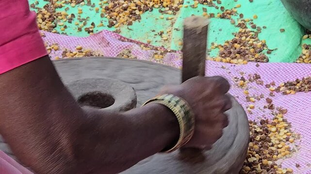 A person operates a traditional hand-cranked stone mill ("chakki") to grind chickpeas/corn. Fingers clear stuck grains from the central hole, maintaining the manual grinding process.