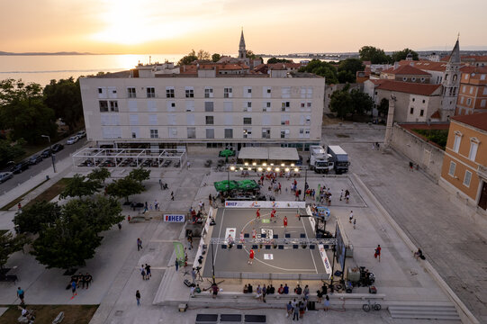 Zadar, Croatia - 18 November 2025: Aerial view of a vibrant basketball game in the city square, framed by historic buildings and the warm glow of the setting sun over the Adriatic Sea. - Powered by Adobe