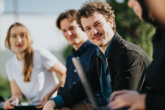A group of young professionals sits outside, discussing work with laptops and notebooks. Friendly smiles and collaborative energy highlight teamwork in a relaxed, outdoor setting.