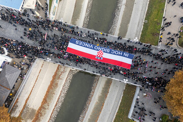 Vukovar, Croatia - 18 November 2025: Aerial view of a solemn procession carrying a Croatian flag banner over a bridge, framed by the city's autumn hues and the flowing Vuka River.