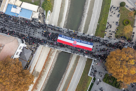 Vukovar, Croatia - 18 November 2025: Aerial view of a solemn procession crossing a bridge, bearing a large Croatian flag emblazoned with 'IN HOC SIGNO VINCES'.