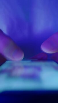 Close-up of hands holding a smartphone and quickly scrolling through social media feeds at night in neon light. Doom scrolling, phone overuse and digital addiction concept. Vertical video