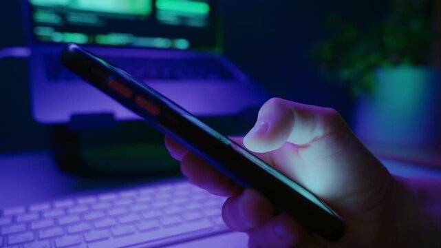 Close-up of persons hand scrolling on smartphone social media feed at night with laptop on background, illuminated by neon light. Digital addiction and modern online habits.