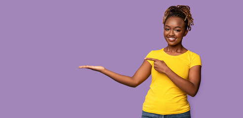 Cheerful african lady pointing at copy space over purple studio background, panorama, space for...