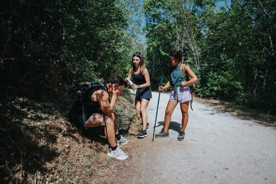 A group of three hikers pauses on a dirt forest path while one friend rests, showing support and teamwork during an outdoor trekking day.