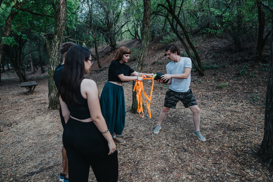 A group of friends in a wooded park engage in a playful outdoor activity, passing bright orange ribbons between hands as they practice teamwork and cooperation amid trees and fallen leaves.