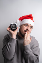 Exhausted man in santa hat yawning holding alarm clock