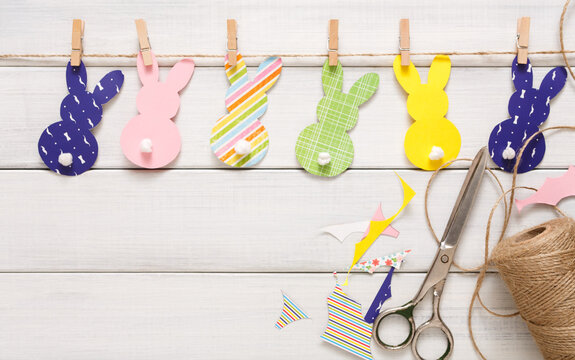 Easter bunny decoration paper cut background. DIY holiday handicraft garland of colorful rabbits and craft tools. Top view with copy space on white wood - Powered by Adobe