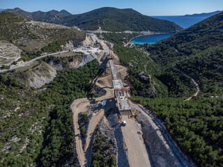 Aerial view of a construction site carving through the landscape, juxtaposing raw earth with the verdant hills and azure waters beyond, Ston, Dubrovnik-Neretva County, Croatia.