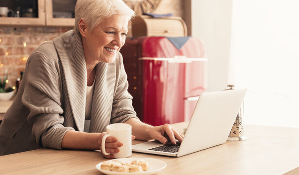 Smiling Aged Woman Relaxing In Kitchen With Laptop And Tea, Browsing Social Media Or Reading News Online, Free Space