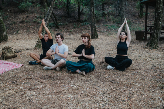 A diverse group of four people sit cross-legged on the forest floor, hands in prayer, enjoying a peaceful outdoor yoga and mindfulness session among trees.
