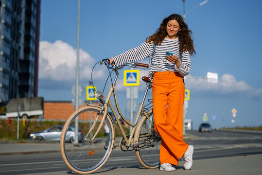 Young adult woman stopping on a city street, checking messages on her smartphone while standing next to her retro bicycle, blending urban mobility with modern communication