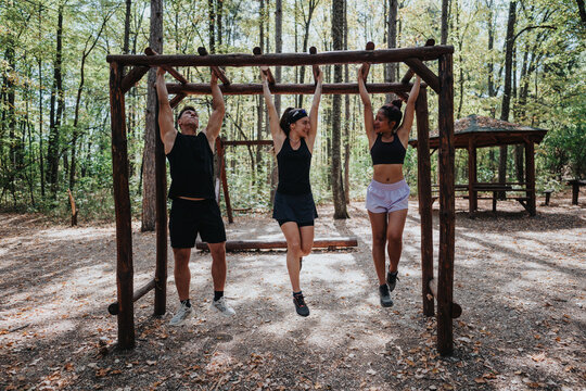 Friends exercise together in a wooded park, hanging from a rustic wooden pull-up frame to strengthen upper body. Bright day and natural surroundings create a lively, healthy outdoor workout scene. - Powered by Adobe