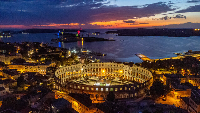 Aerial view of the illuminated Pula Arena glows against the twilight sky, surrounded by the city's lights and the Adriatic Sea, Pula, Istria County, Croatia.