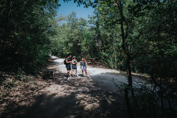 Obraz premium A small group hike on a sunny forest trail, equipped with backpacks and trekking poles, sharing a moment outdoors near a wooden bench amid lush trees.