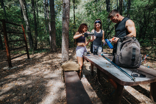 A group of friends enjoys a sunny outdoor break in the woods, pouring coffee, organizing gear, and sharing a moment near a rustic picnic table while surrounded by trees.