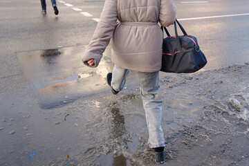 Woman jumps over slushy puddle while crossing winter road, challenge of navigating slippery pedestrian areas during cold weather and highlighting seasonal urban walking hazards