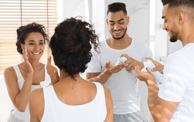 Morning Rituals. Happy Arab Couple Making Self-Care Routine Near Mirror In Bathroom, Middle Eastern Lovers Getting Ready Together, Man Using Shaving Foam While Woman Applying Moisturising Cream