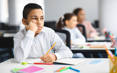 Education And Learning Concept. Portrait of tired and bored small boy sitting at desk in classroom at school, writing in notebook and thinking, looking away at window, resting head on hand © Prostock-studio