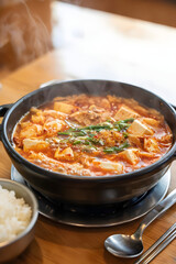 Authentic Kimchi Jjigae with Soft Tofu and Pork on Wooden Table - Steaming Spicy Korean Stew in Cozy Restaurant with White Rice and Warm Bokeh Background
