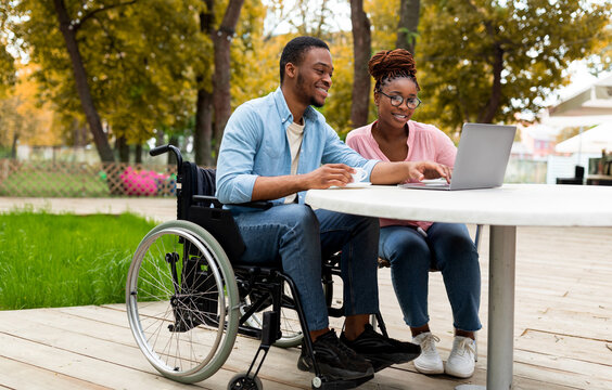 Cheerful African American man in wheelchair with woman colleague using laptop together in outdoor cafe at autumn park, working on online business project, attending meeting remotely