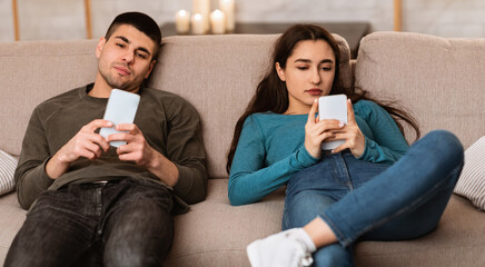 Boredom Concept. Young couple using smart phones, sitting on couch at home during self isolation