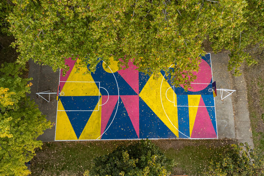 Aerial view of a vibrant basketball court painted with geometric shapes in bold pink, yellow, and blue, nestled amongst verdant trees, Osijek, Osijek-Baranja County, Croatia.