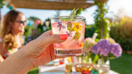 Hand holding Refreshing floral cocktail outdoors at summer garden party setting. Refreshingly vibrant floral ice drink held in garden setting for summer gatherings.