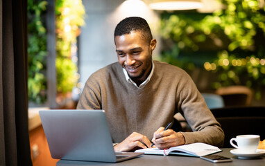 Inspired African American manager working in cafe, using laptop and taking notes in his organizer, empty space