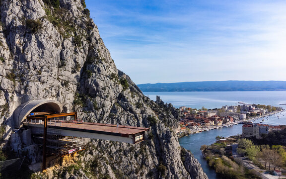 Aerial view of a modern viewing platform extending from a craggy, sunlit cliffside over a shimmering river towards the historic town of Omis, Split-Dalmatia County, Croatia.
