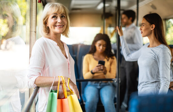 Public Transportation. Group of diverse people taking bus, selective focus on smiling mature woman holding shopping bags, standing and looking out of window, leaning on handle going home
