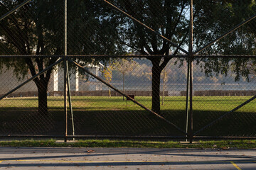 Fence, field, park, trees, sunlight, outdoors scene with chain-link mesh and boundary in foreground; quiet open ground beyond shade, grass, and clear sky.