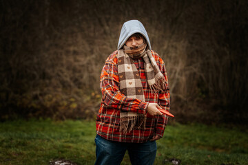 A boy in a red plaid jacket and scarf throws a red discus in a grassy park with trees. Concentrated outdoor activity in cool weather.