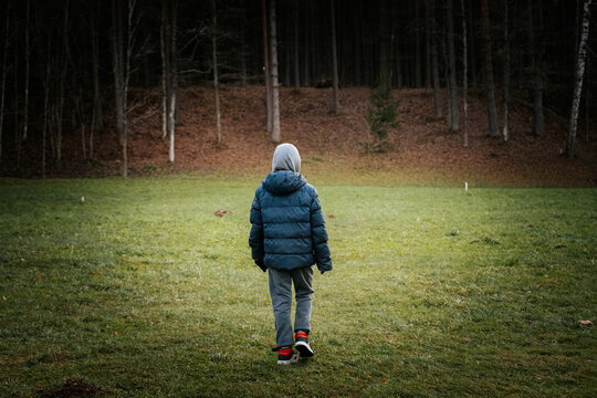 A child in a hooded winter coat walks across a grassy clearing toward a dense forest. Overcast, earthy tones create a calm and solitary mood.