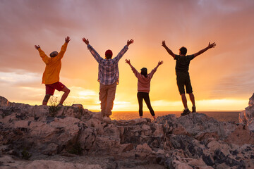 Four happy diverse tourists are having fun and meeting sunset with open arms at lake shore against colorful pink and orange cloudy sky. Active vacations concept