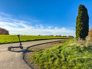 Fototapeta premium Electric scooter parked on a winding country road beside a tall cypress, fresh green grass and wide blue sky. Peaceful countryside scene in Luxembourg.