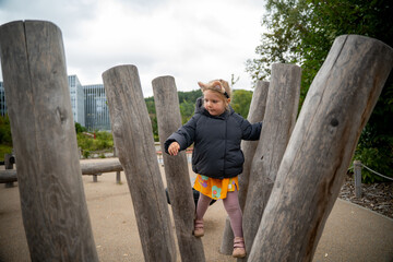 A little girl wearing a cat ear headband and black puffer jacket explores a wooden obstacle course at an eco-friendly playground in a park on a cold autumn day. Childhood curiosity, outdoor activity.